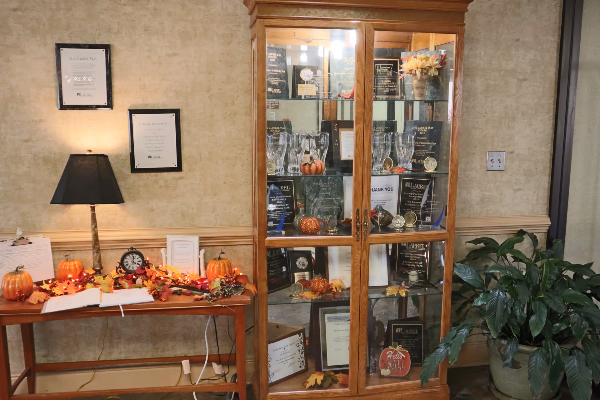 A decorated interior area featuring a glass-front trophy cabinet, a table with a lamp and fall decorations, and a potted plant.