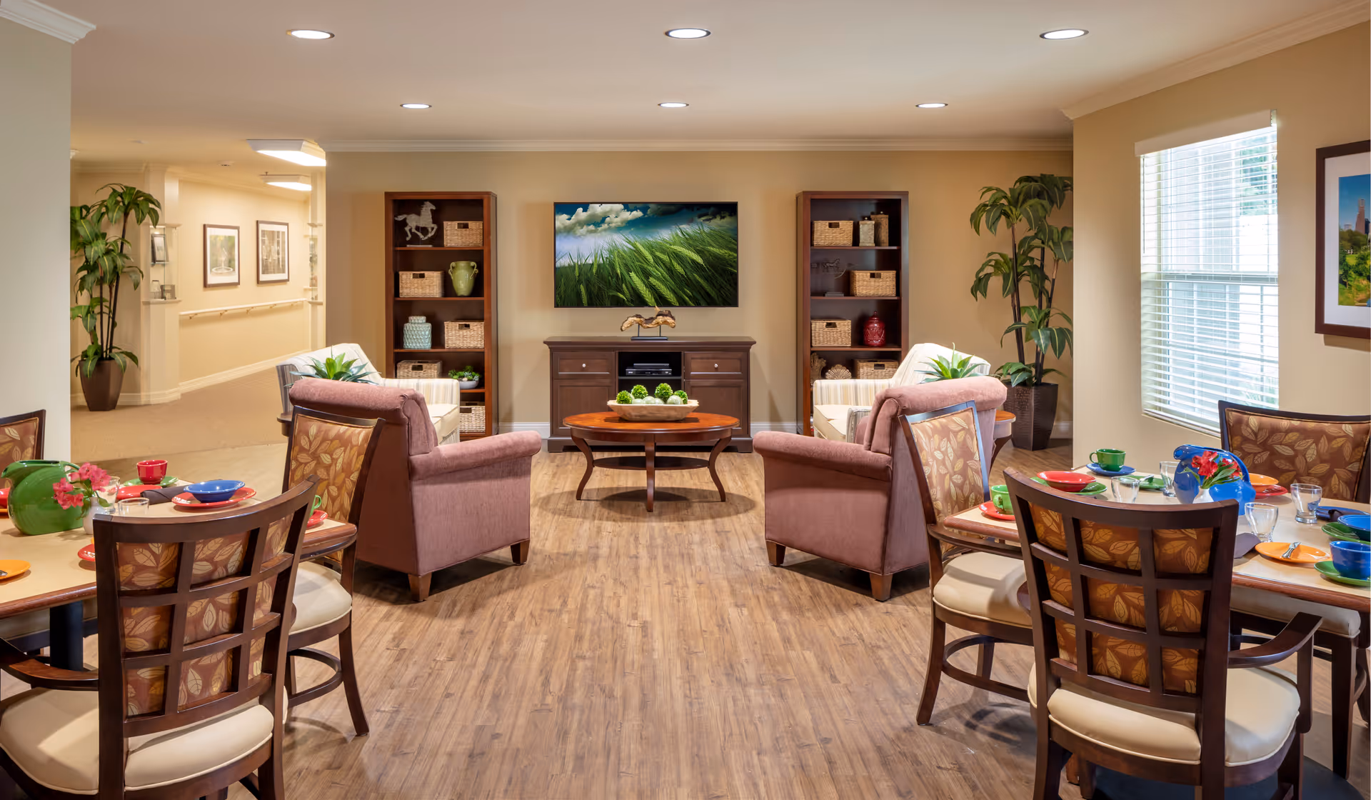 A cozy senior living common area with two armchairs facing a flat-screen TV mounted on the wall. The room features wooden flooring, two tall bookshelves flanking the TV stand, and two dining tables with chairs set with colorful dishes and cups. Large windows allow natural light to brighten the space, and potted plants add greenery to the room.