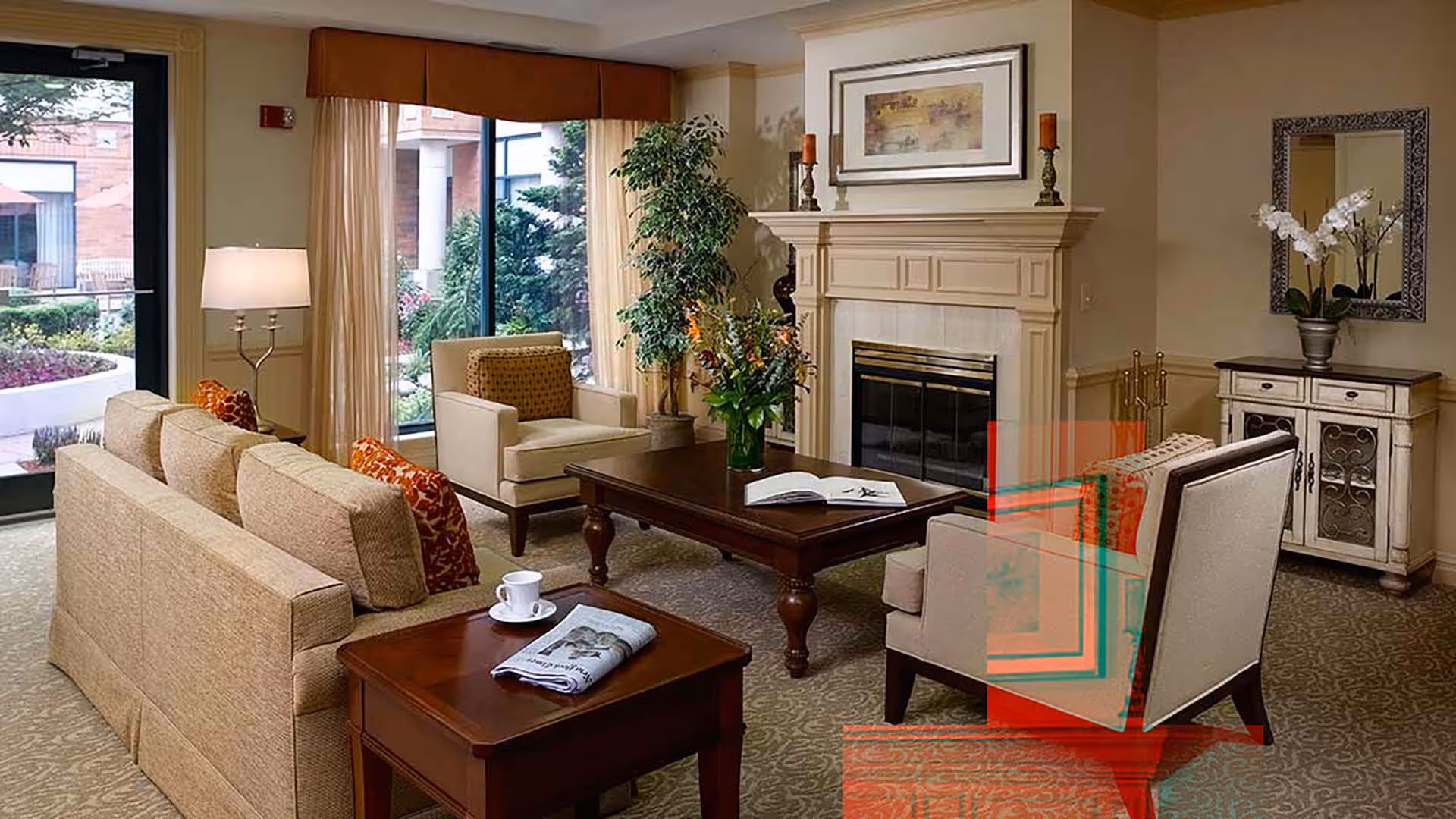 A cozy living room area with beige sofas and armchairs arranged around a dark wooden coffee table with a vase of flowers and an open book. There is a side table with a cup and a folded newspaper. The room features a fireplace with decorative candles and a framed picture above it. Large windows with curtains provide a view of a garden outside. A small cabinet with a mirror and a potted orchid is against the wall.