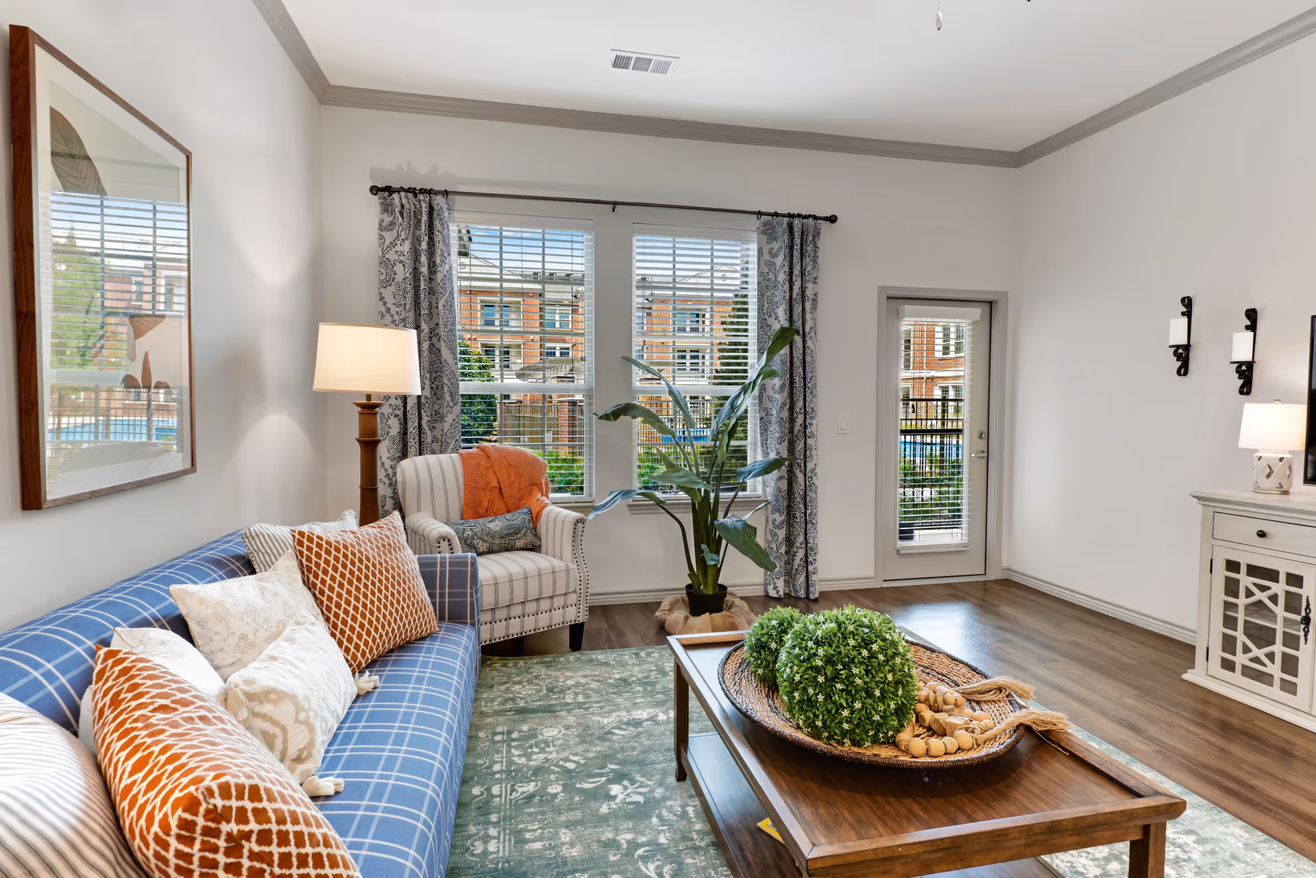 Bright staged living room with a blue plaid sofa, armchair, coffee table with decorative tray, and large windows and door to the outside.