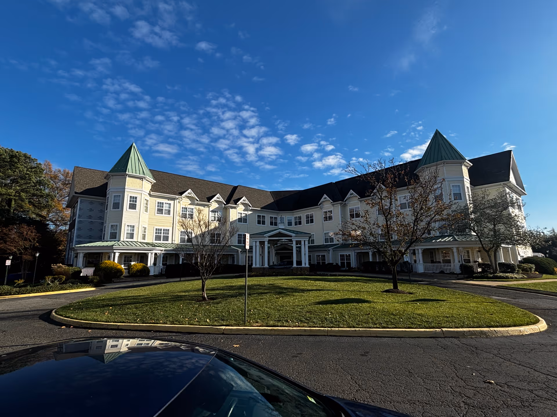 Exterior view of a large, three-story senior living facility building with green peaked roofs and multiple windows under a blue sky with scattered clouds. There is a circular driveway with a grassy island and a few small trees in front of the building.