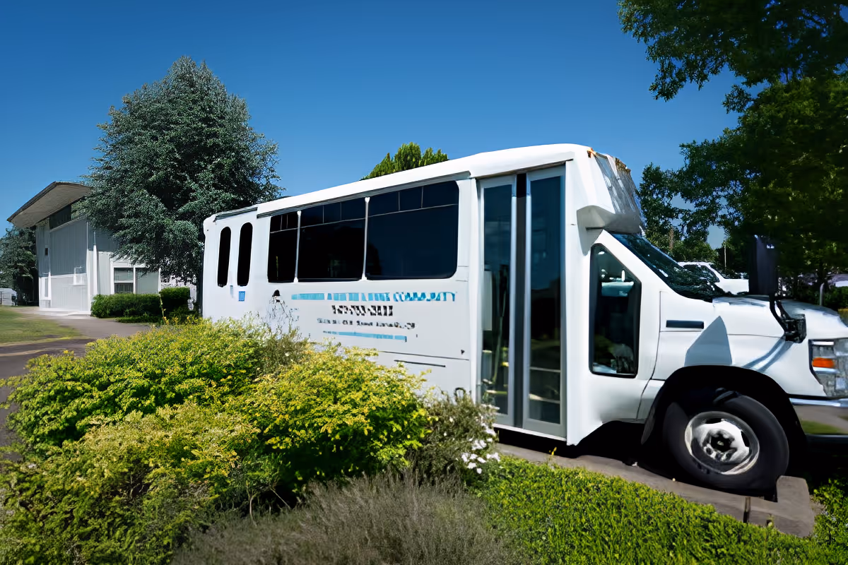 A white shuttle bus parked on a driveway next to green bushes and trees under a clear blue sky, with a building visible in the background.