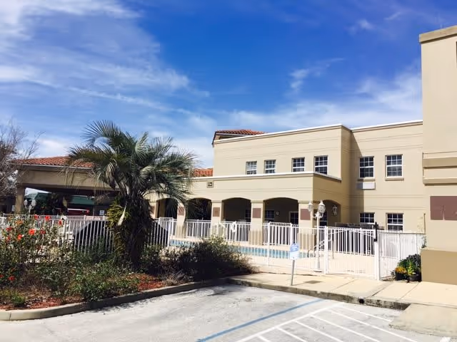 Exterior view of a beige two-story building with multiple windows and a red-tiled roof under a blue sky. In front of the building is a fenced area with a swimming pool, surrounded by some palm trees and shrubs. A parking area with marked spaces is visible in the foreground.
