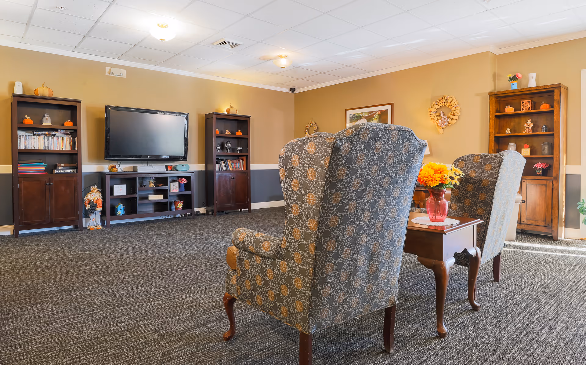 A cozy living room area in an assisted living facility featuring two patterned armchairs facing a flat-screen TV mounted on the wall. The room has beige walls with a gray lower trim, carpeted floor, and wooden bookshelves decorated with small pumpkins and other ornaments. A small wooden side table with a vase of yellow and orange flowers is placed between the chairs.