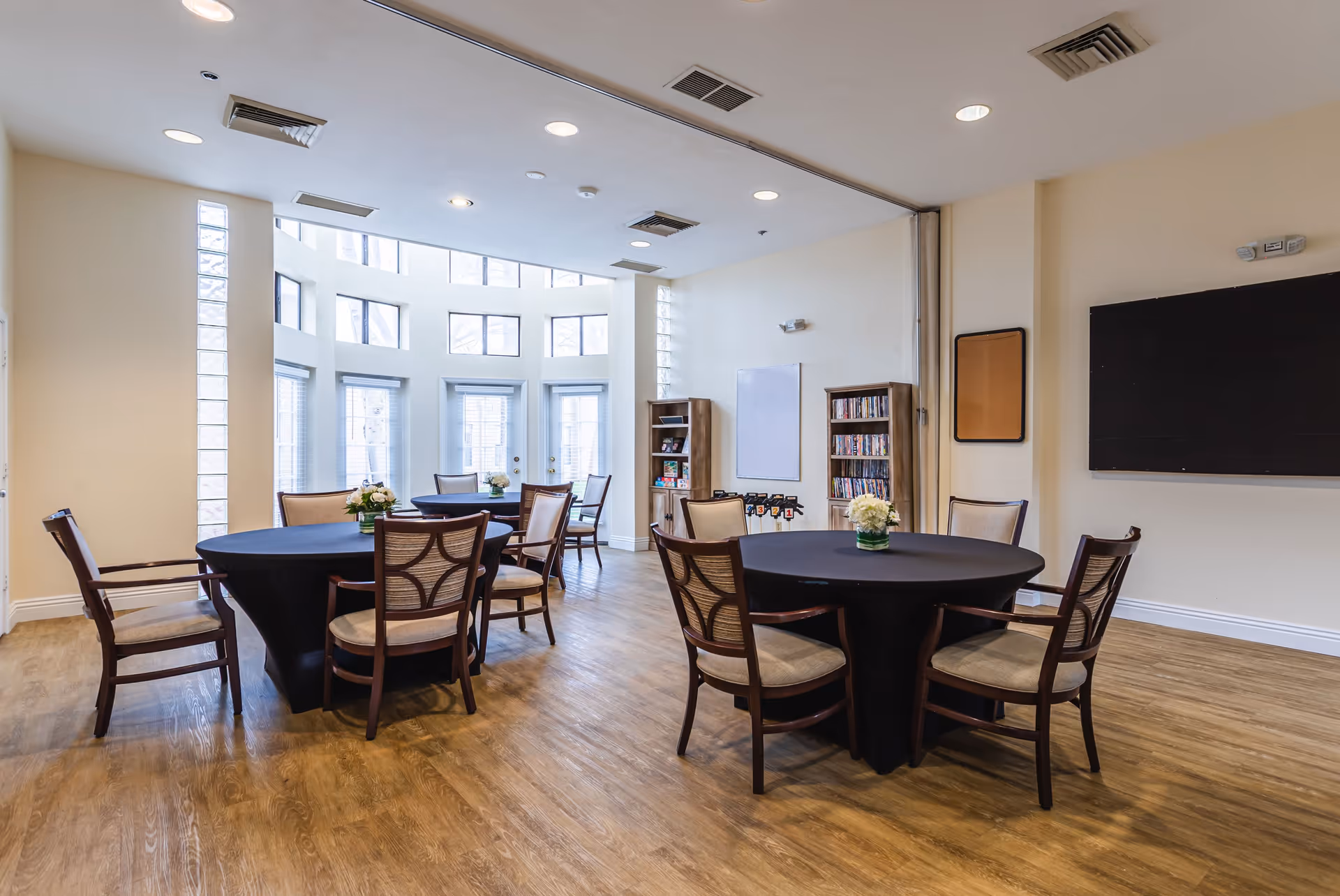 A bright and spacious common area with round tables covered in black tablecloths, each surrounded by wooden chairs with beige cushions. The room features large windows allowing natural light to fill the space, wooden flooring, and bookshelves against the walls. There is a large blackboard or screen mounted on one wall.
