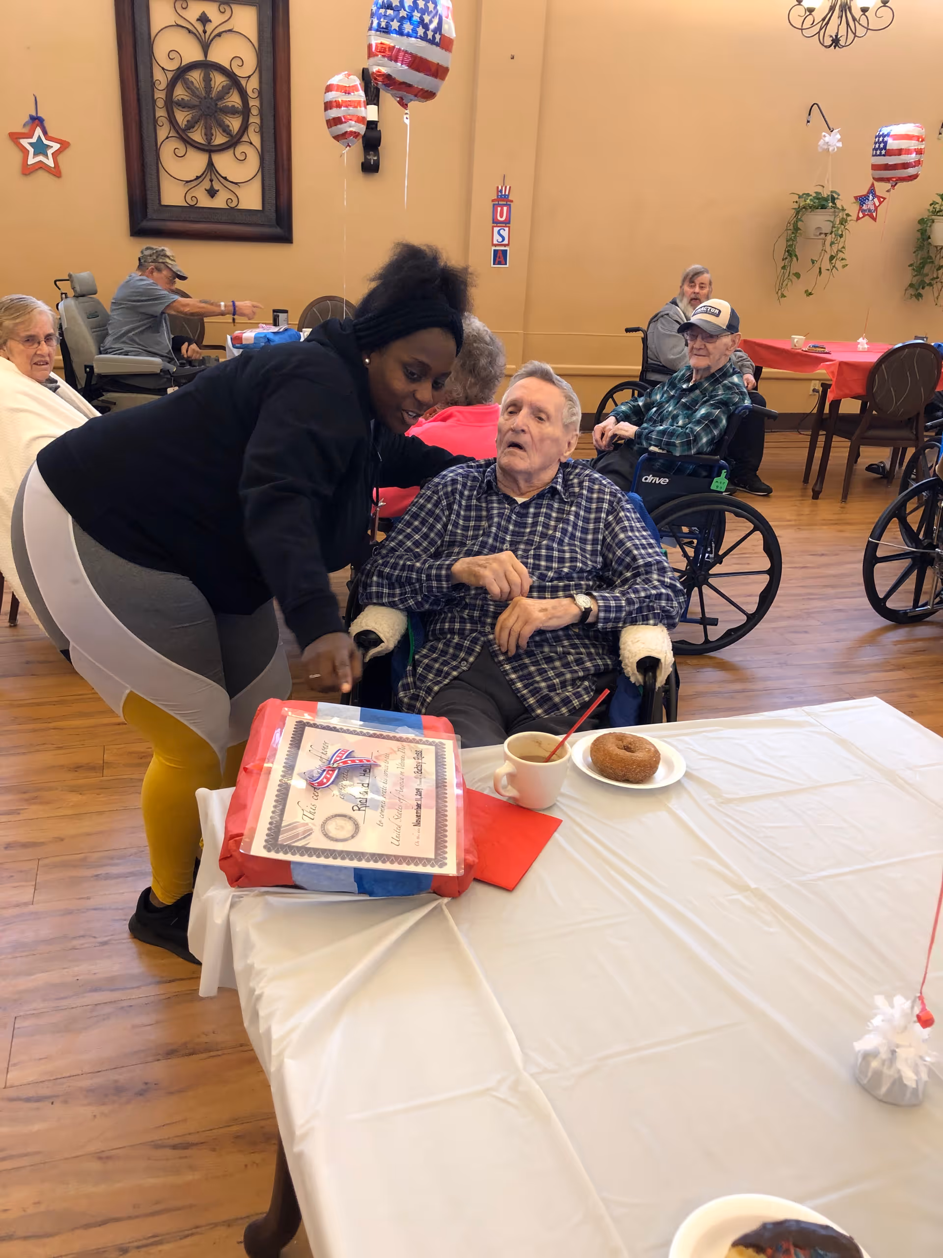 A caregiver leans over an elderly man in a wheelchair at a decorated dining table while other residents in wheelchairs sit in the background.