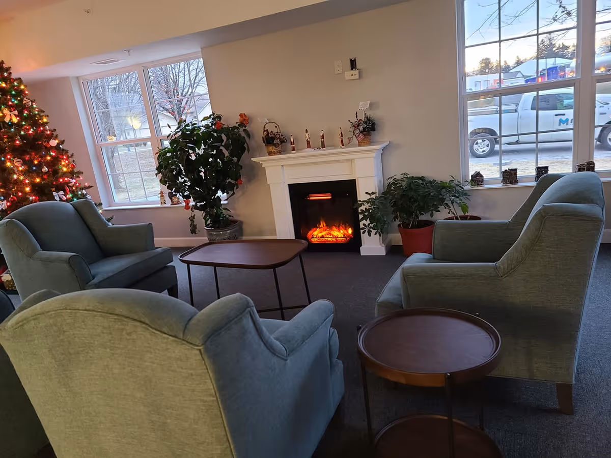 A cozy sitting area with four green upholstered armchairs arranged around two small wooden tables. There is a white electric fireplace with decorative items on the mantel, two large potted plants on either side, and a decorated Christmas tree with colorful lights near a window. Outside the windows, a white pickup truck and some buildings are visible.