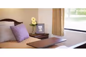 A neatly made bedroom with a bed, pillows, a bedside table holding flowers and a framed photo, a tray table across the bed, and a window with curtains.