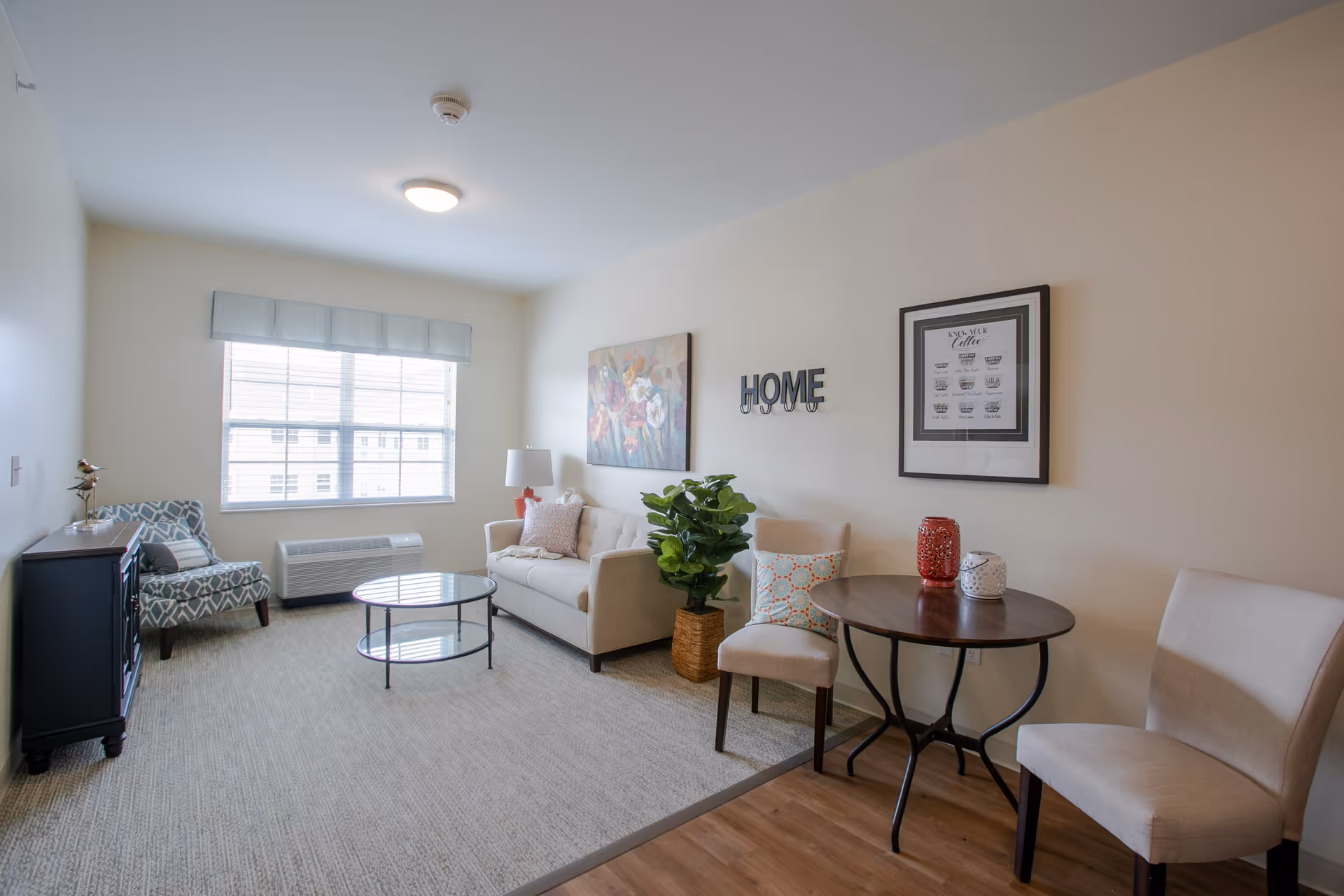 Bright furnished living room with a sofa, armchairs, round coffee table and small dining table beneath wall art that says 'HOME' and a large window.