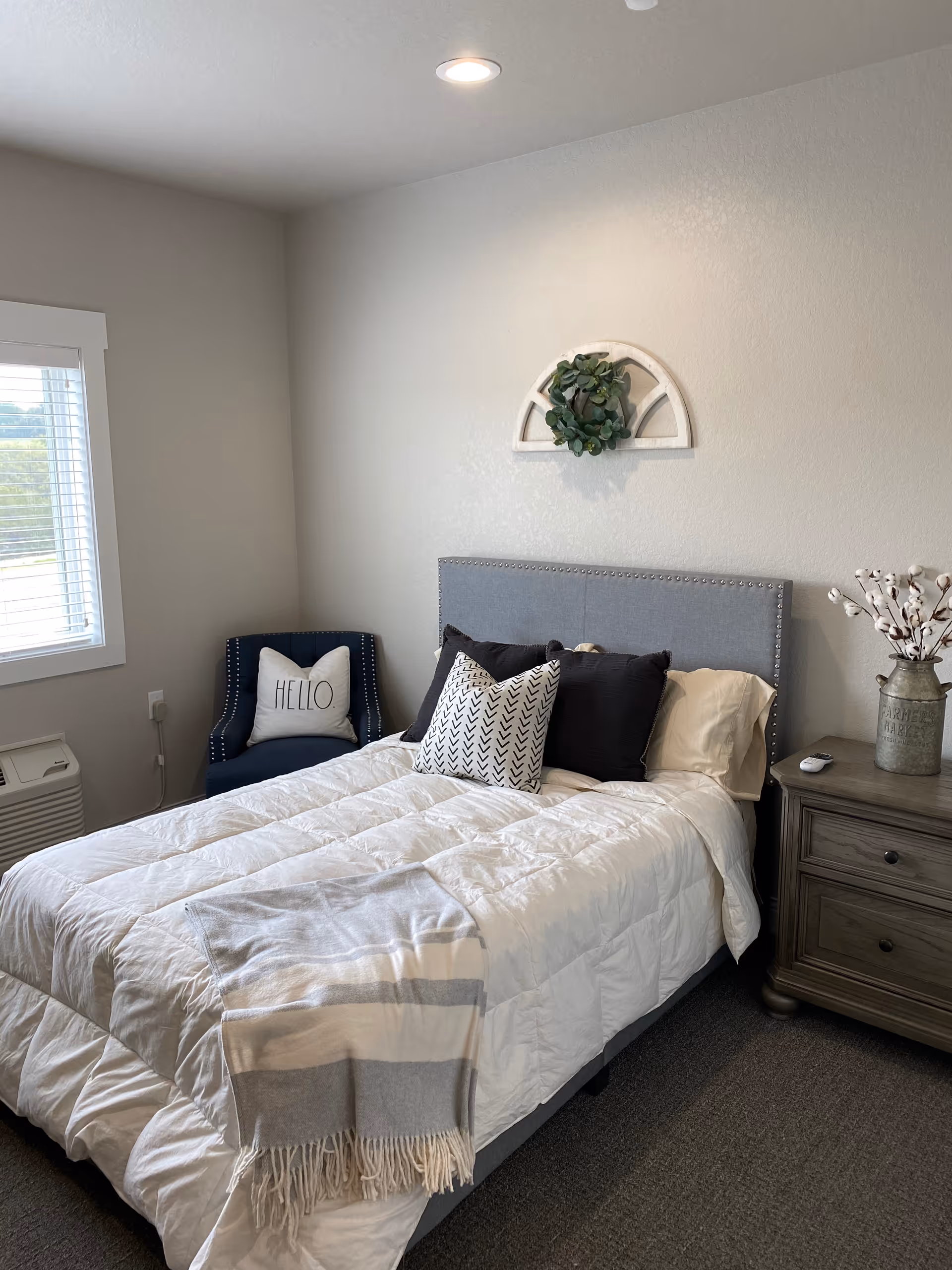 A cozy bedroom with a neatly made bed featuring a gray upholstered headboard, white comforter, and decorative pillows. A gray and white striped throw blanket is draped over the foot of the bed. To the right is a wooden nightstand with a vase of cotton stems and a remote control. In the corner near a window with white blinds, there is a dark blue armchair with a white pillow that says 'HELLO'. Above the bed, a decorative half-circle wall hanging with greenery is mounted on the light gray wall.