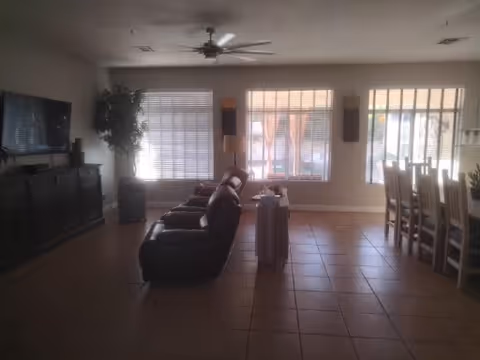 Interior view of a living room with a row of three dark recliner chairs facing a large flat-screen TV mounted on a dark wooden cabinet. There is a large potted plant near a window with blinds, and a ceiling fan above. To the right, there is a dining area with a wooden dining table and chairs near large windows with blinds and a sliding glass door.