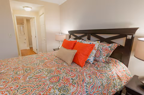 A bedroom with a large bed featuring a colorful paisley-patterned bedspread and multiple pillows in red and beige. There are two bedside tables with lamps on either side of the bed. In the background, an open door reveals a bathroom with a shower and wooden flooring.