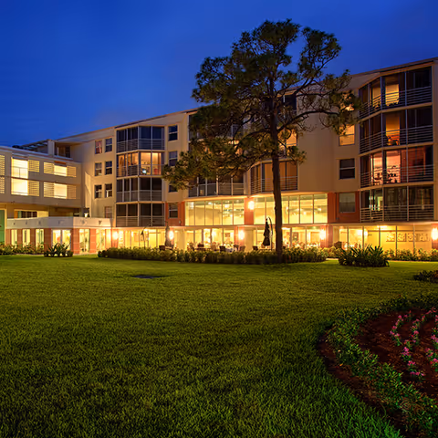 Exterior view of a multi-story senior living facility at dusk with well-maintained green lawn and a large tree in front. The building has large windows with warm interior lighting visible, and landscaped flower beds near the lawn.