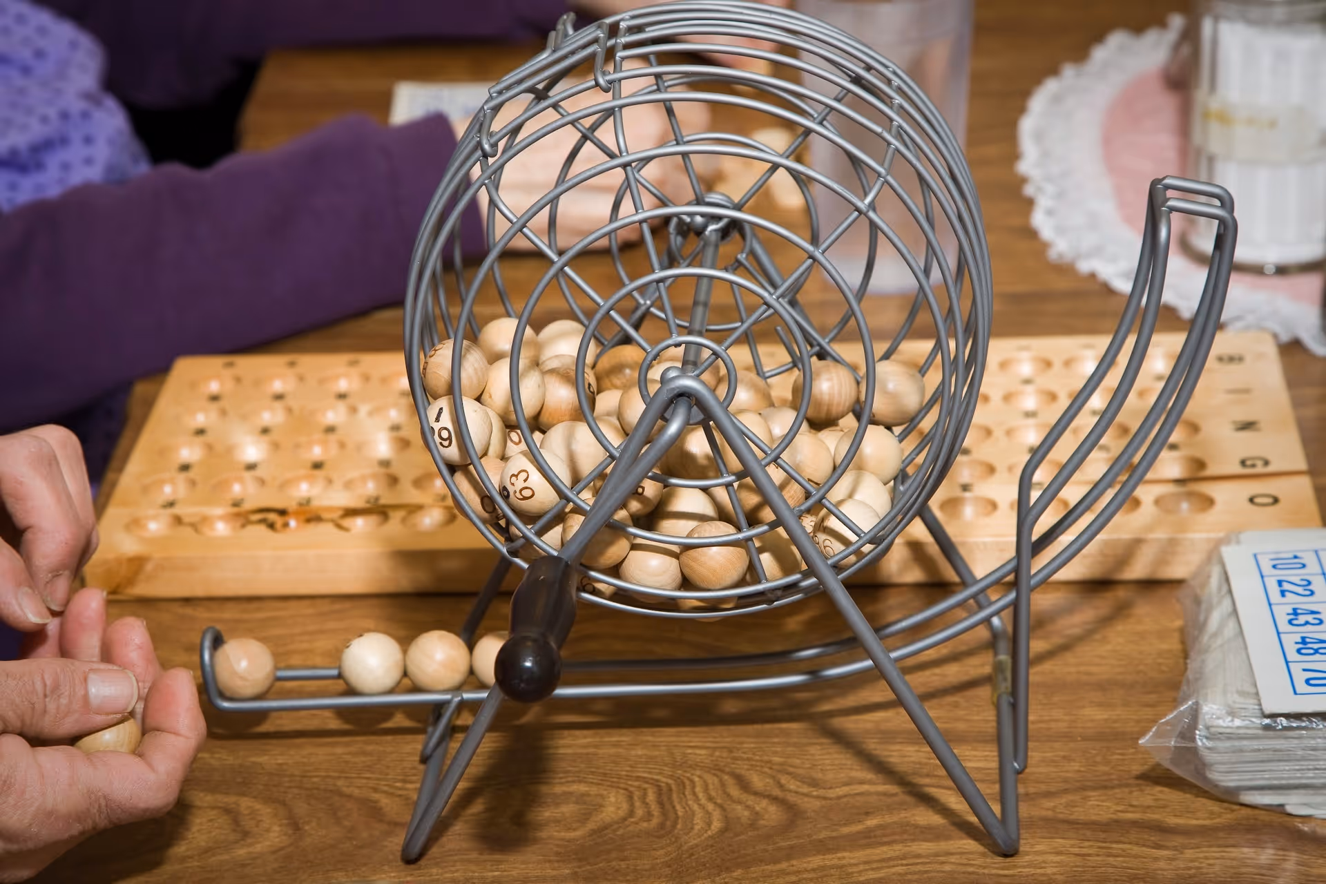 Close-up of a bingo game setup on a wooden table with a metal cage containing numbered wooden balls. Two hands are visible, one holding a ball and the other resting on the table near a bingo board. A stack of bingo cards and a glass with a doily underneath are also on the table.