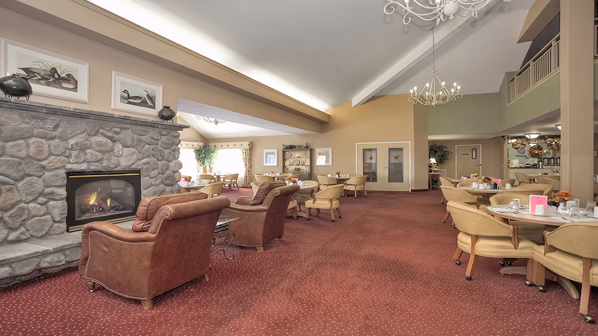 Spacious communal dining and lounge area with a stone fireplace, leather armchairs, round dining tables, chandeliers, and red carpet.