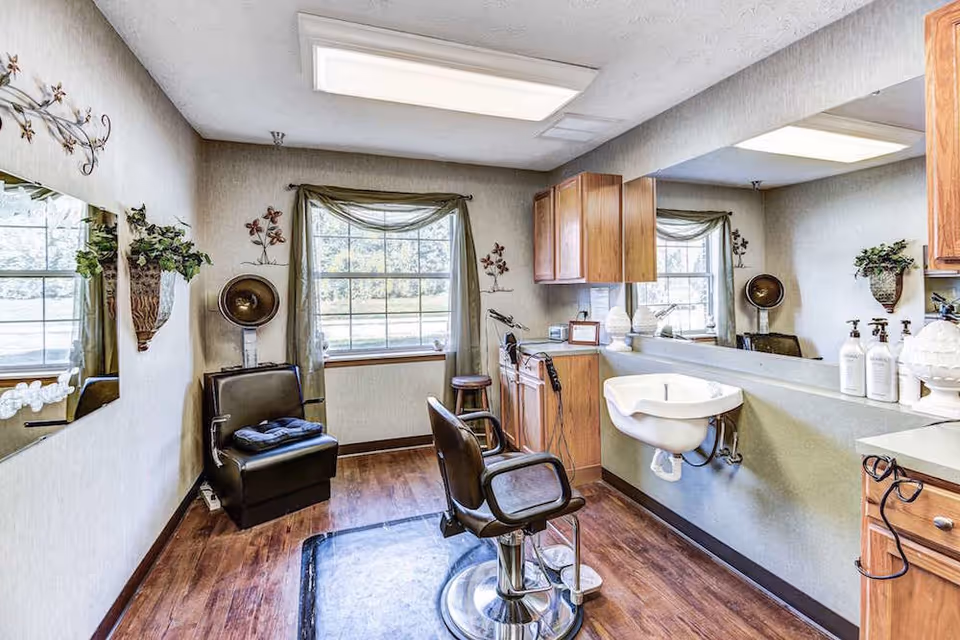 Interior view of a salon area in a senior living facility with a black salon chair, a hair dryer chair, a large mirror on the wall, wooden cabinets, a sink, and a window with green curtains letting in natural light.