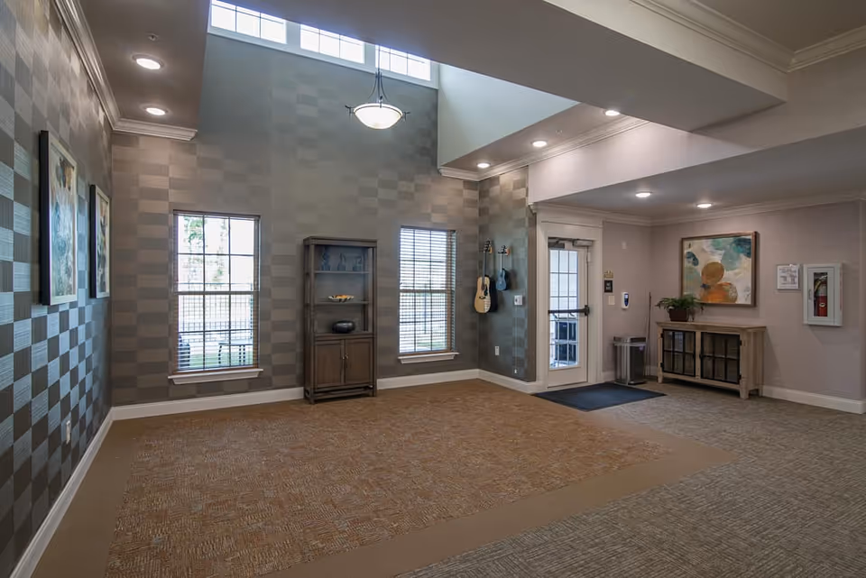 An interior common area with high ceilings and large windows letting in natural light. The walls have a checkered pattern wallpaper, and there are two guitars hanging on one wall. A wooden cabinet with shelves and a sideboard with a plant and artwork above it are also visible. The floor is carpeted with two different patterns, and there is a glass door leading outside.