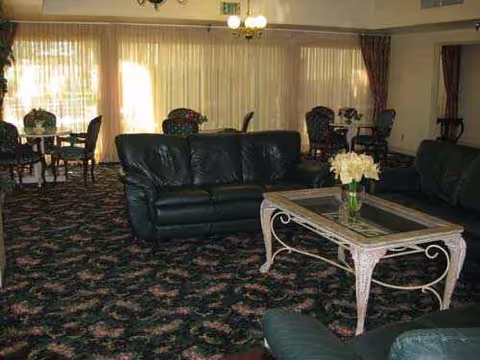 Senior living lounge with dark leather sofas, floral carpet, a glass coffee table, and sheer curtains.