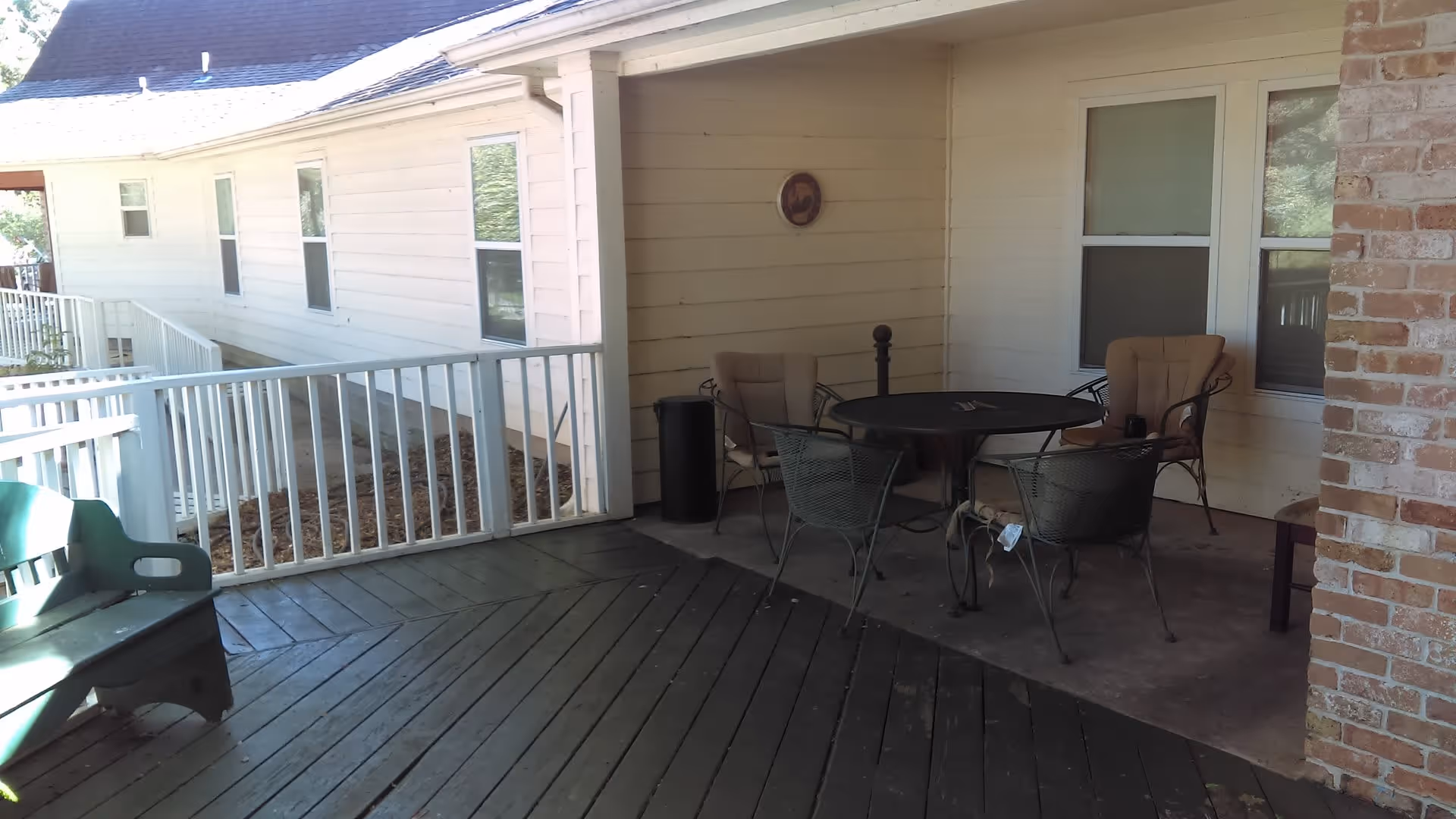 Outdoor covered patio area with a round metal table and four chairs, two with cushions. The patio has a wooden floor and white railing, with a brick column on the right side and windows on the wall behind the table.