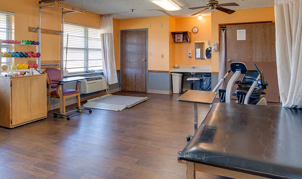 Spacious interior therapy room with treatment tables, exercise equipment, hand weights, chairs, and large windows.