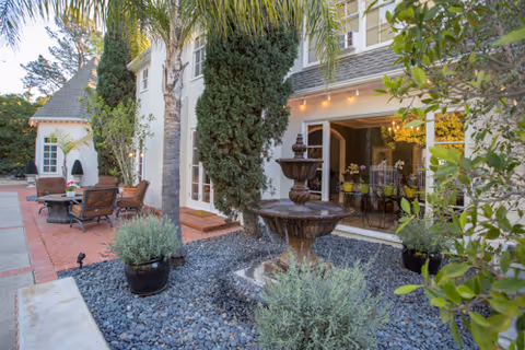Outdoor patio area at Chateau Rose featuring a multi-tiered water fountain surrounded by plants and trees, with a seating area including chairs and tables on a brick patio. The building exterior is visible with large windows and a door leading inside, where a dining area with a chandelier can be seen.