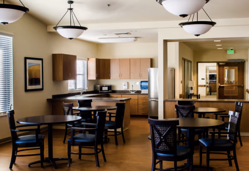 A dining area with several round tables and chairs, adjacent to a kitchen with wooden cabinets, a refrigerator, a microwave, and a sink. The room has beige walls, large windows with blinds, ceiling lights, and a hallway leading to another room.