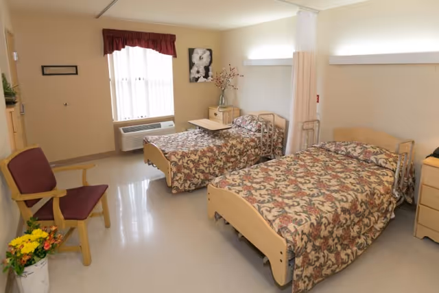 A clean and well-lit room with two single beds covered in floral patterned bedspreads. There is a wooden chair with a maroon cushion, a small table with a vase of flowers, and a window with vertical blinds and a red valance. The room has beige walls and a light-colored floor.