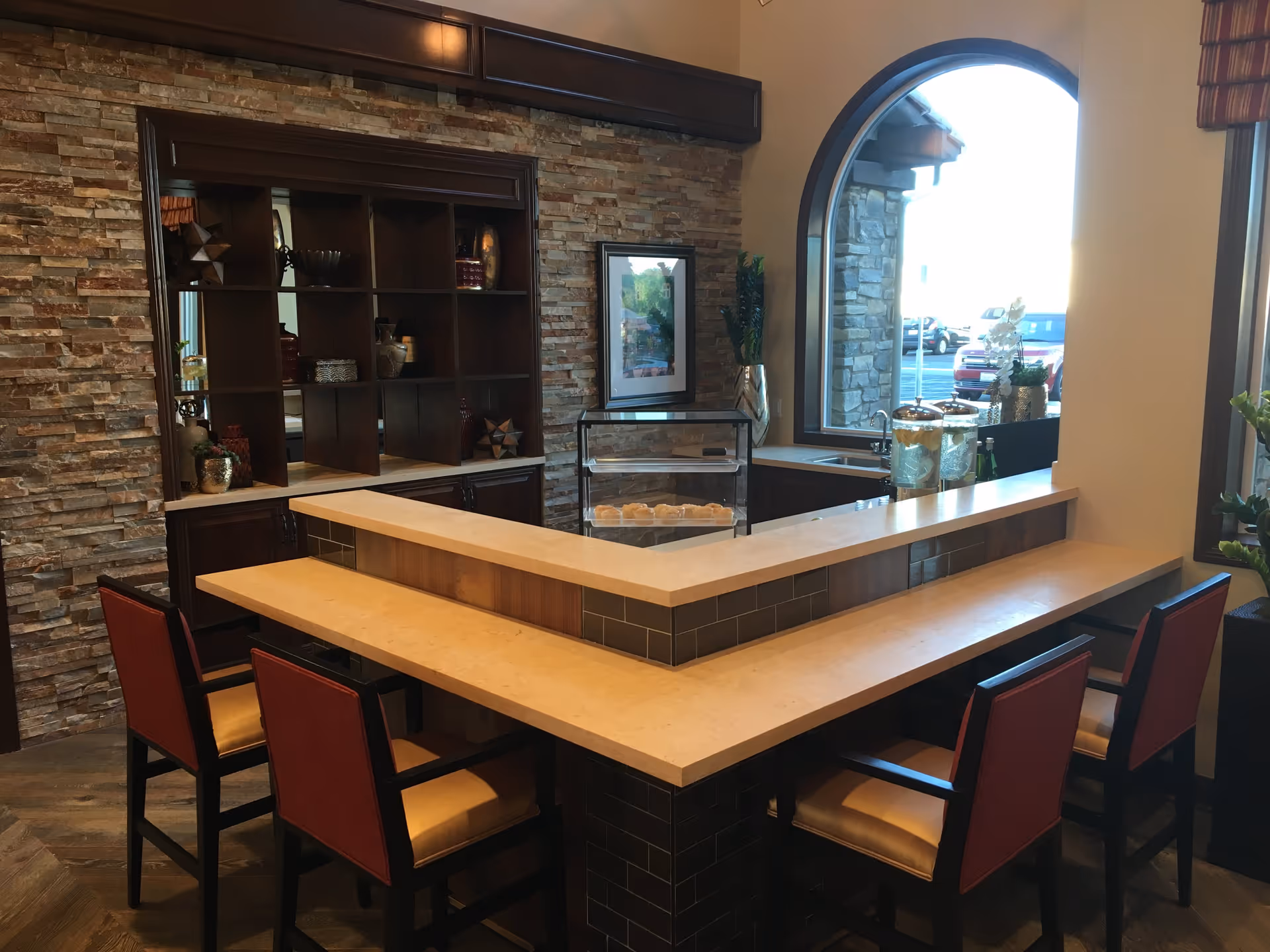 Interior view of a seating area with a U-shaped counter and four chairs with red backs and beige seats. The counter is made of light-colored stone with a dark tiled base. Behind the counter is a stone accent wall with dark wooden shelves displaying decorative items. A large arched window lets in natural light and shows a parking lot outside.