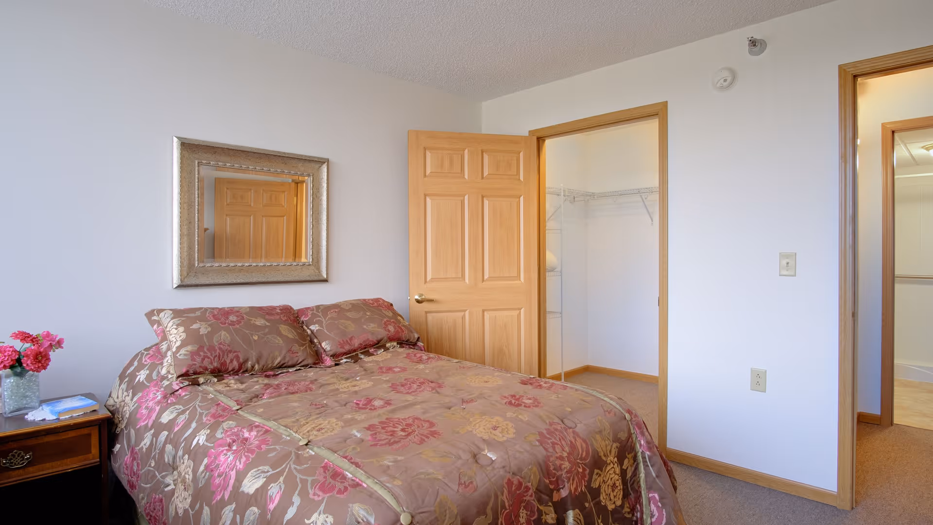 A bedroom with a bed covered in a floral patterned comforter in shades of pink and beige. There is a wooden nightstand with a vase of pink flowers and some books. A large mirror with a silver frame hangs on the wall above the bed. The room has light-colored walls and carpeted floor. An open wooden door reveals a walk-in closet with shelving and hanging rods. Another open doorway leads to a hallway with a glimpse of a bathroom.