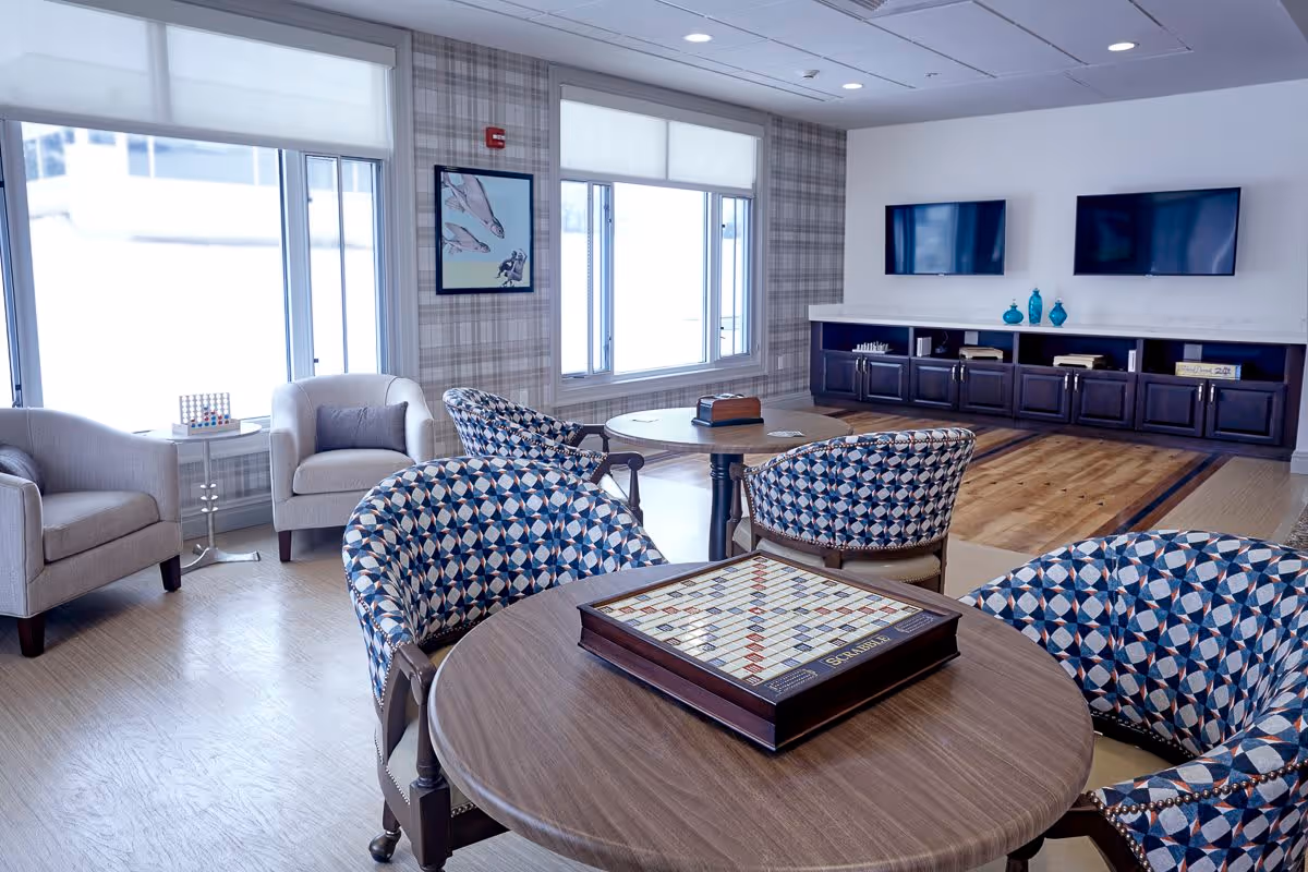 A bright and cozy common area with patterned armchairs around wooden tables, one table featuring a Scrabble game set. The room has large windows with white blinds, plaid wallpaper, two wall-mounted flat-screen TVs above a dark wooden cabinet, and light wood flooring.
