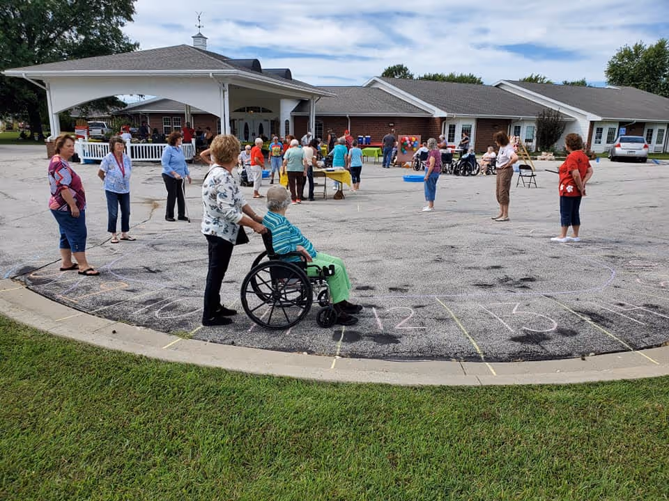 Residents and staff gathered outside a senior care building for an activity in a parking lot with chalk markings, including a person in a wheelchair.