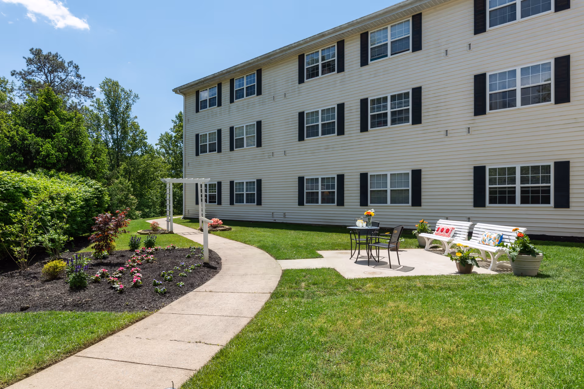 Outdoor patio area at Washington Township Senior Living with a concrete walkway, a small garden bed with flowers and shrubs, a white pergola, a black metal table with two chairs, and two white benches with colorful cushions on a grassy lawn beside a three-story building with multiple windows and black shutters.