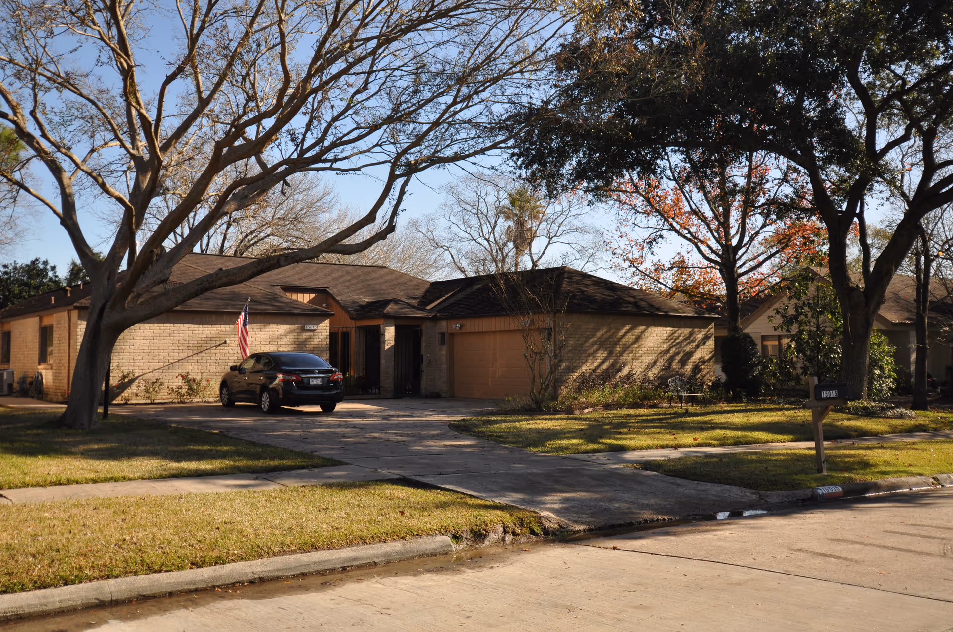 Front view of a single-story brick house with a driveway, a parked car, large trees, and an American flag.