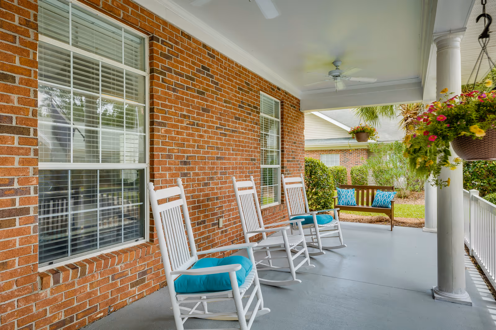 A covered porch area with a brick wall and two large windows. The porch features three white rocking chairs with blue cushions and a wooden bench with blue patterned pillows. There are hanging flower baskets and white columns supporting the roof, with ceiling fans overhead. The porch overlooks a green lawn and shrubbery.