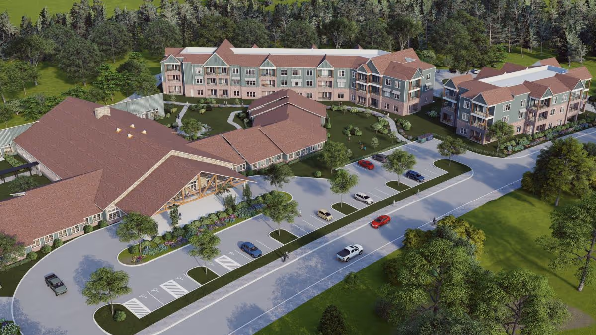Aerial view of The Timberley senior living facility showing multiple connected buildings with red roofs surrounded by trees and greenery. There is a parking lot with several cars and a road in front of the buildings.
