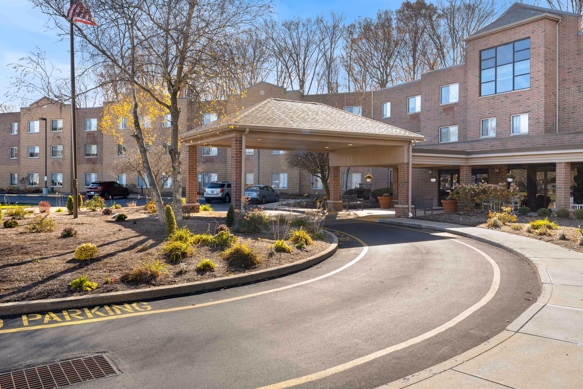Exterior view of Whispering Oaks - A Provincial Senior Living Community, showing a curved driveway leading to a covered entrance with brick buildings in the background, landscaped garden beds, and an American flag on a flagpole.