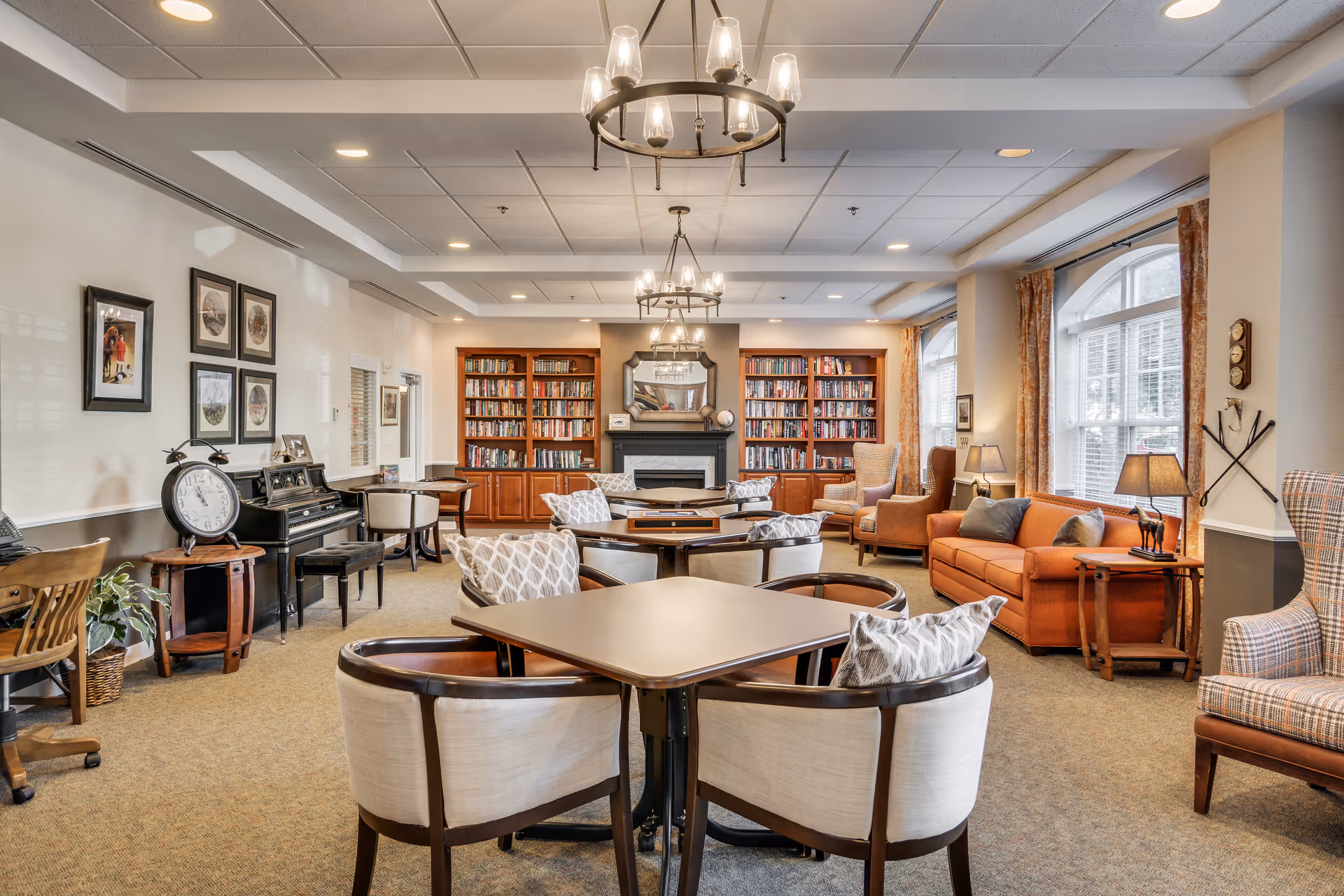 A cozy and well-lit living room area in an assisted living facility featuring multiple seating arrangements with cushioned chairs and sofas, a fireplace flanked by built-in bookshelves filled with books, large windows with curtains allowing natural light, a piano with a large clock on a side table, framed pictures on the wall, and decorative lighting fixtures hanging from the ceiling.