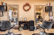 Interior view of a salon area with two styling chairs facing large mirrors mounted on a beige wall. Between the mirrors is a small black cabinet with hair styling tools and products. A decorative wreath hangs on the wall above the cabinet. Shelves with additional hair products and framed pictures are visible on either side of the mirrors.