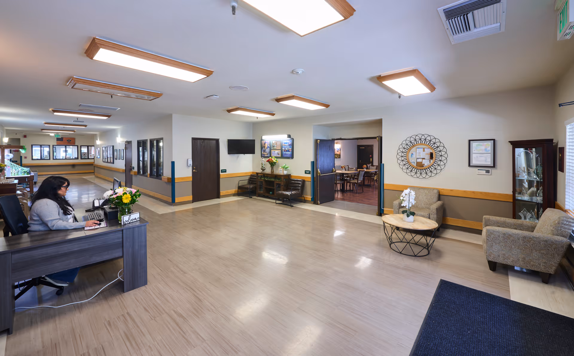 A spacious and well-lit reception area of a nursing and rehabilitation center with a woman sitting at a desk on the left side working on a computer. The room features light-colored flooring, beige walls with a wooden trim, ceiling lights, a small seating area with two armchairs and a round table with a white orchid, a wall-mounted TV, and a hallway leading to other rooms.