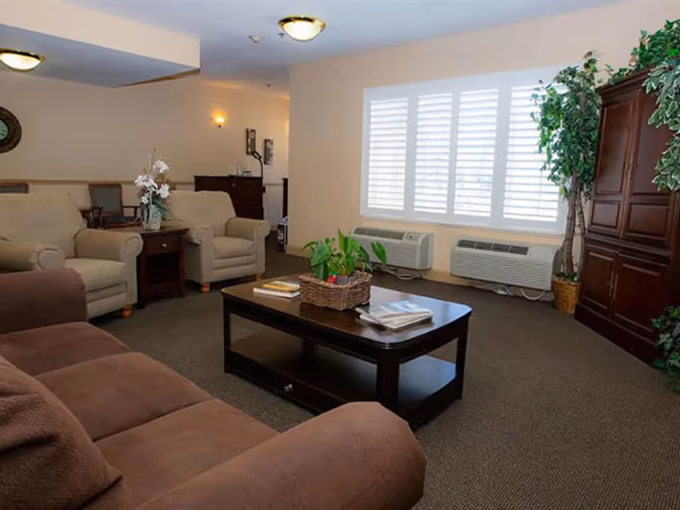 A cozy living room with a brown sofa, two beige armchairs, a dark wood coffee table with books and a plant basket, large window with white blinds, air conditioning units below the window, and a wooden cabinet with green plants on either side.
