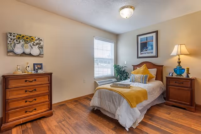 A cozy bedroom with a single bed dressed in white and yellow bedding, a wooden headboard, and a bedside table with a blue lamp and a candle. There is a wooden dresser with decorative items and a framed picture on the wall above it. A window with blinds lets in natural light, and a potted plant is placed near the window. The room has wooden flooring and neutral-colored walls.