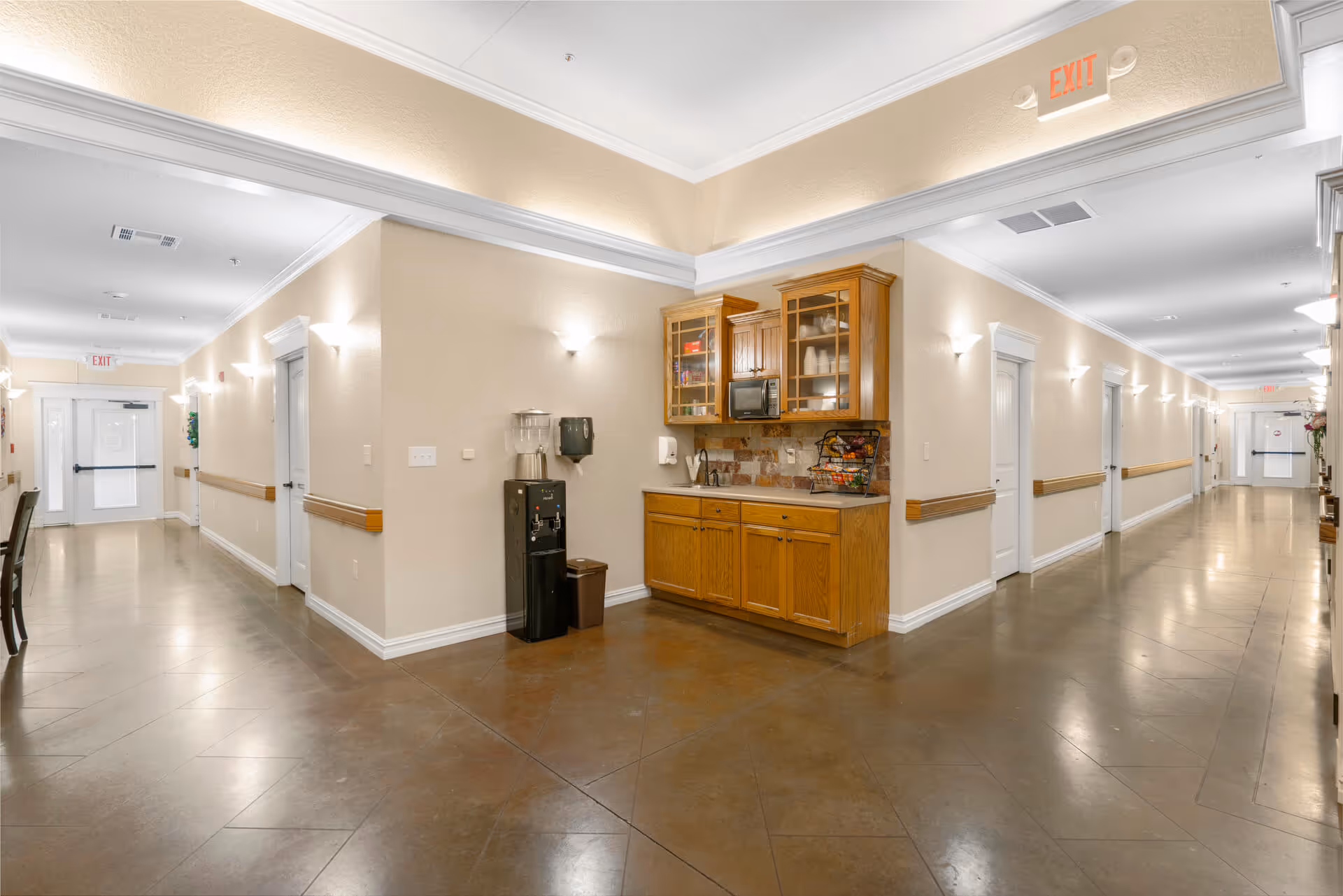 A well-lit hallway in a senior living facility with beige walls, brown tiled floors, and white trim. The hallway features multiple white doors along both sides and exit signs at the ends. In the center corner, there is a small kitchenette area with wooden cabinets, a microwave, a water dispenser, and a basket of fruit.