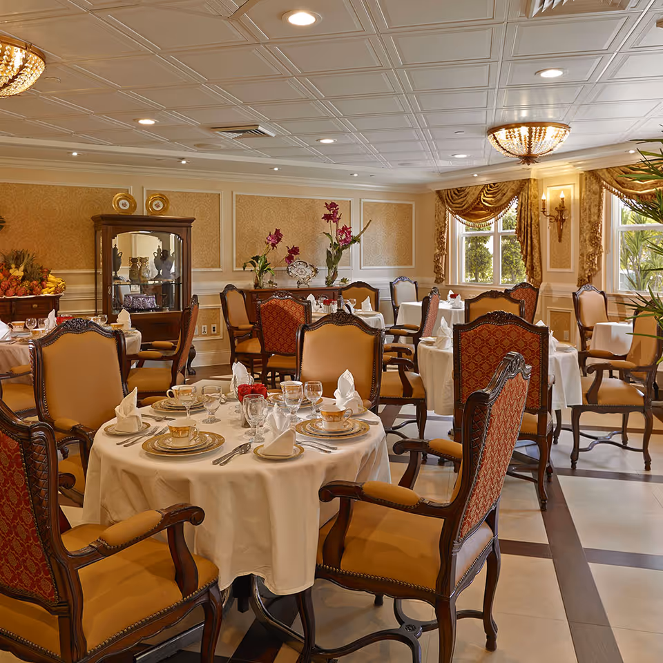 Elegant dining room with round tables covered in white tablecloths, set with fine china, glassware, and folded napkins. The room features ornate wooden chairs with upholstered seats and backs, decorative wall panels, large windows with draped curtains, and floral arrangements on sideboards.