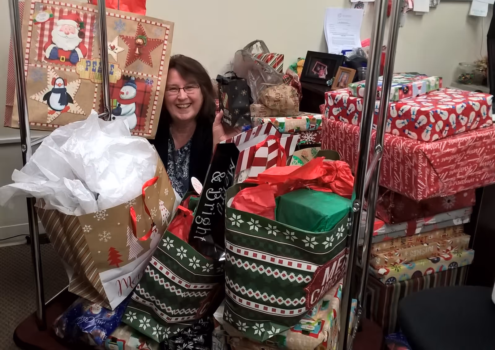 A woman smiling behind a large collection of Christmas gift bags and wrapped presents stacked on a luggage cart in an indoor setting.