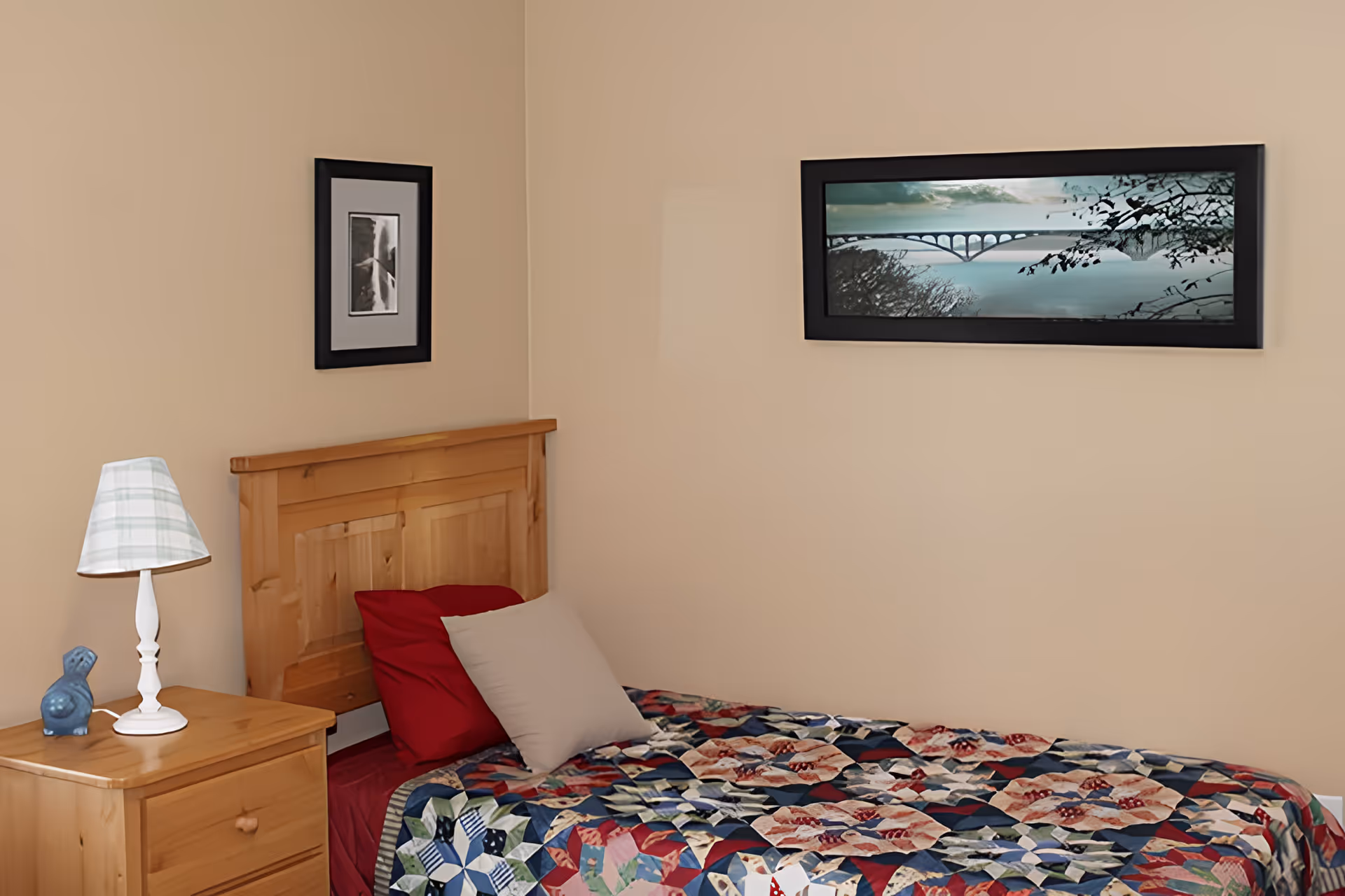 A small bedroom with a single wooden bed featuring a colorful patchwork quilt and two pillows, one red and one beige. Next to the bed is a wooden nightstand with a white lamp with a plaid lampshade and a small blue decorative object. The walls are beige and have two framed pictures hanging, one black and white and one color landscape of a bridge over water.