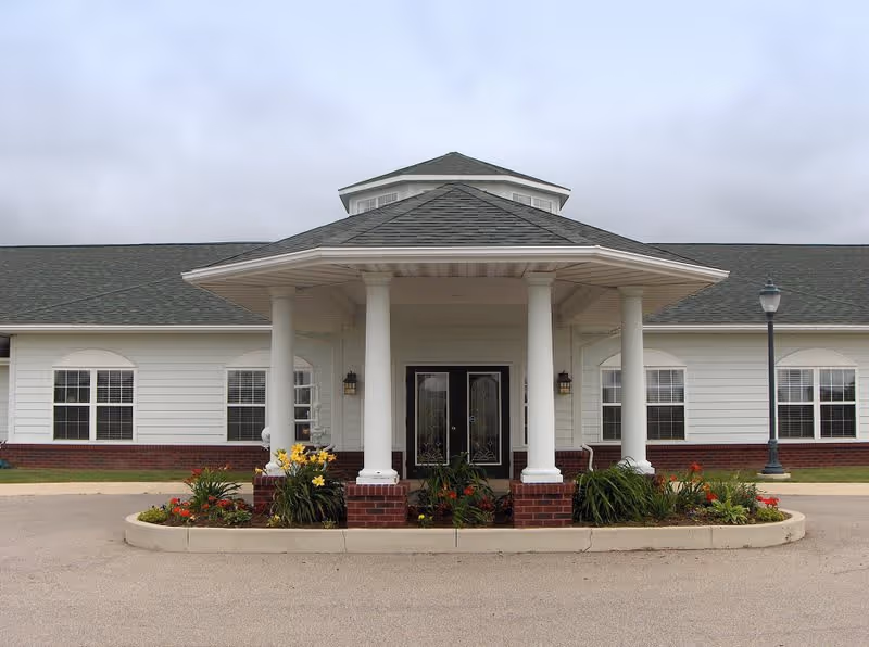 Front exterior view of a single-story building with white siding and a gray shingled roof. The entrance features a covered portico supported by four white columns and a landscaped flower bed with various plants and flowers in front. There are multiple windows with white frames on either side of the entrance, and a street lamp is visible on the right side of the building.
