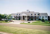 Front exterior view of a large, two-story assisted living facility named Prairie Gardens Assisted Living, featuring a symmetrical design with a central entrance supported by white columns, multiple windows, and a well-maintained lawn and driveway in front.