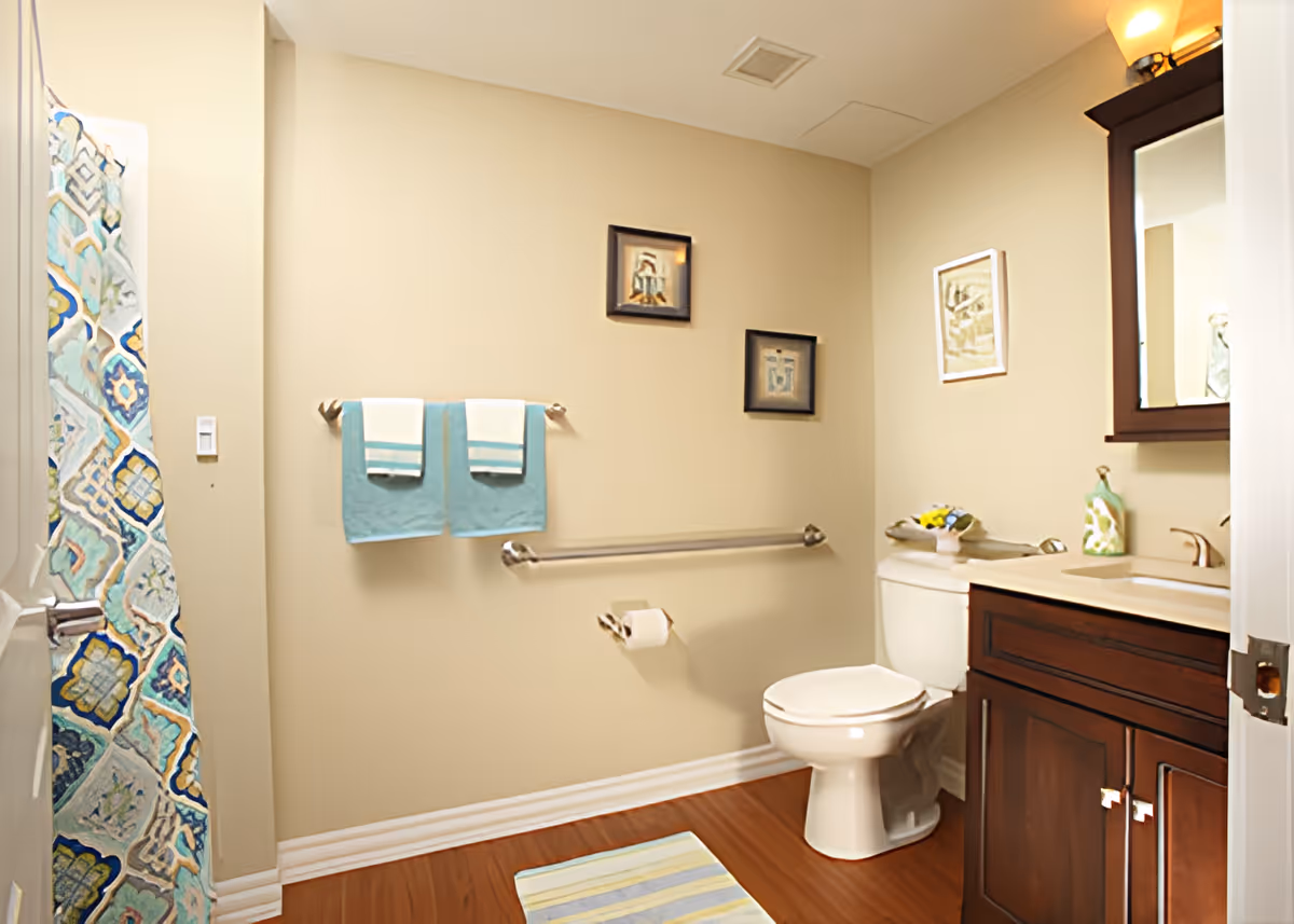 A clean bathroom with beige walls and wooden floor. There is a white toilet next to a dark wooden vanity with a sink and mirror above it. Two blue and white towels hang on a towel rack on the wall, and a colorful patterned shower curtain is partially visible on the left. The bathroom is decorated with framed pictures and a small flower arrangement on the toilet tank.
