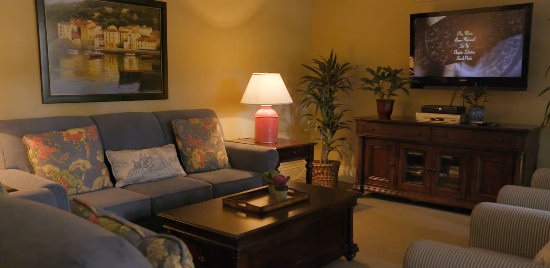 A cozy living room with a gray sofa adorned with floral and patterned cushions, a wooden coffee table with a small plant and tray, a side table with a pink lamp, a large framed painting on the wall, a wooden cabinet with a flat-screen TV mounted above it, and two striped armchairs.