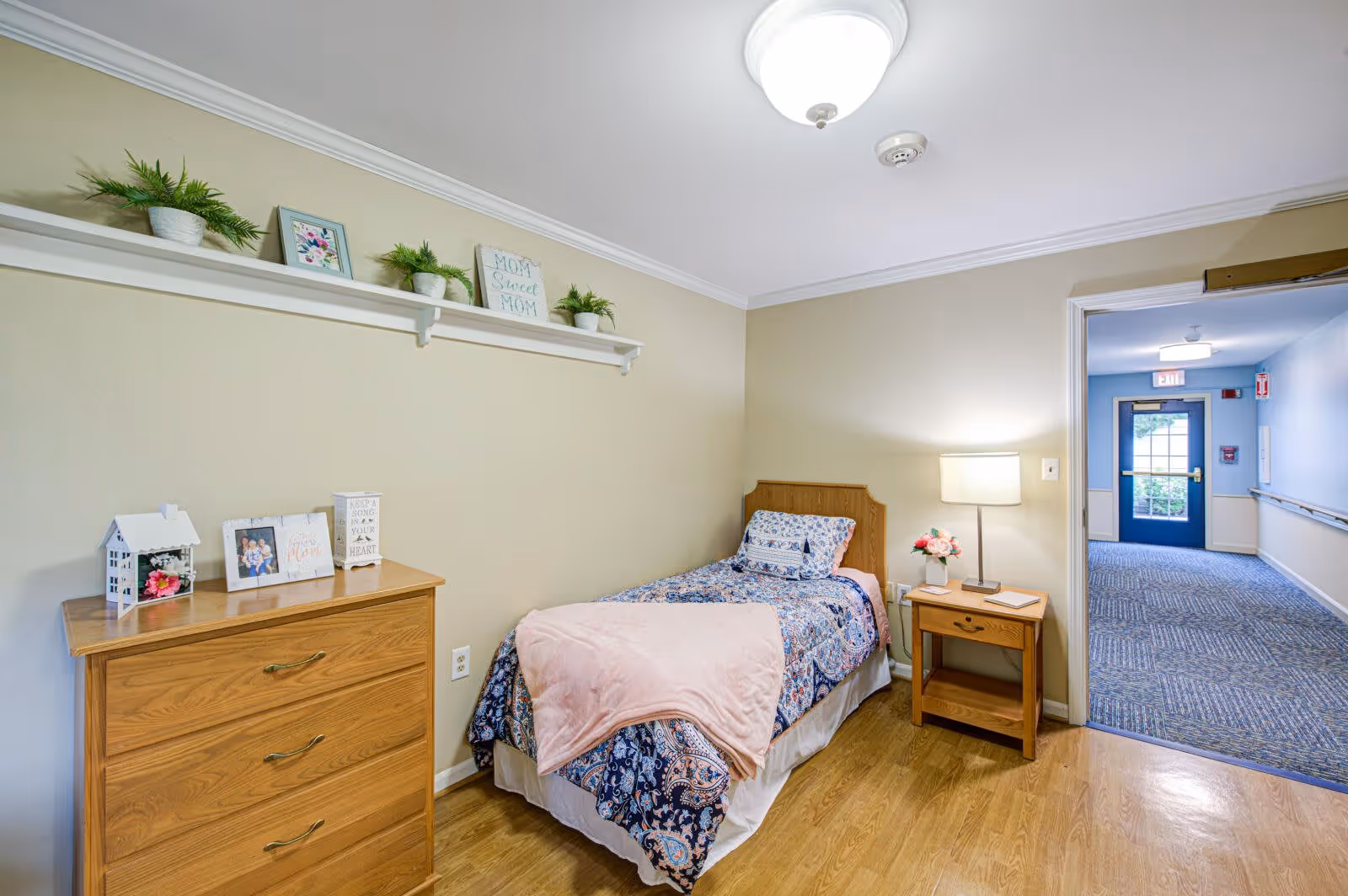 A small bedroom with a single bed covered in a blue and white patterned bedspread and a pink blanket. Next to the bed is a wooden nightstand with a lamp and a small vase of flowers. On the left side of the room is a wooden dresser with framed photos and decorative items on top. Above the dresser is a white shelf with small potted plants and framed pictures. The room has light-colored walls and wood flooring. An open doorway leads to a hallway with blue walls and carpeted floor, and a door with a window at the end.